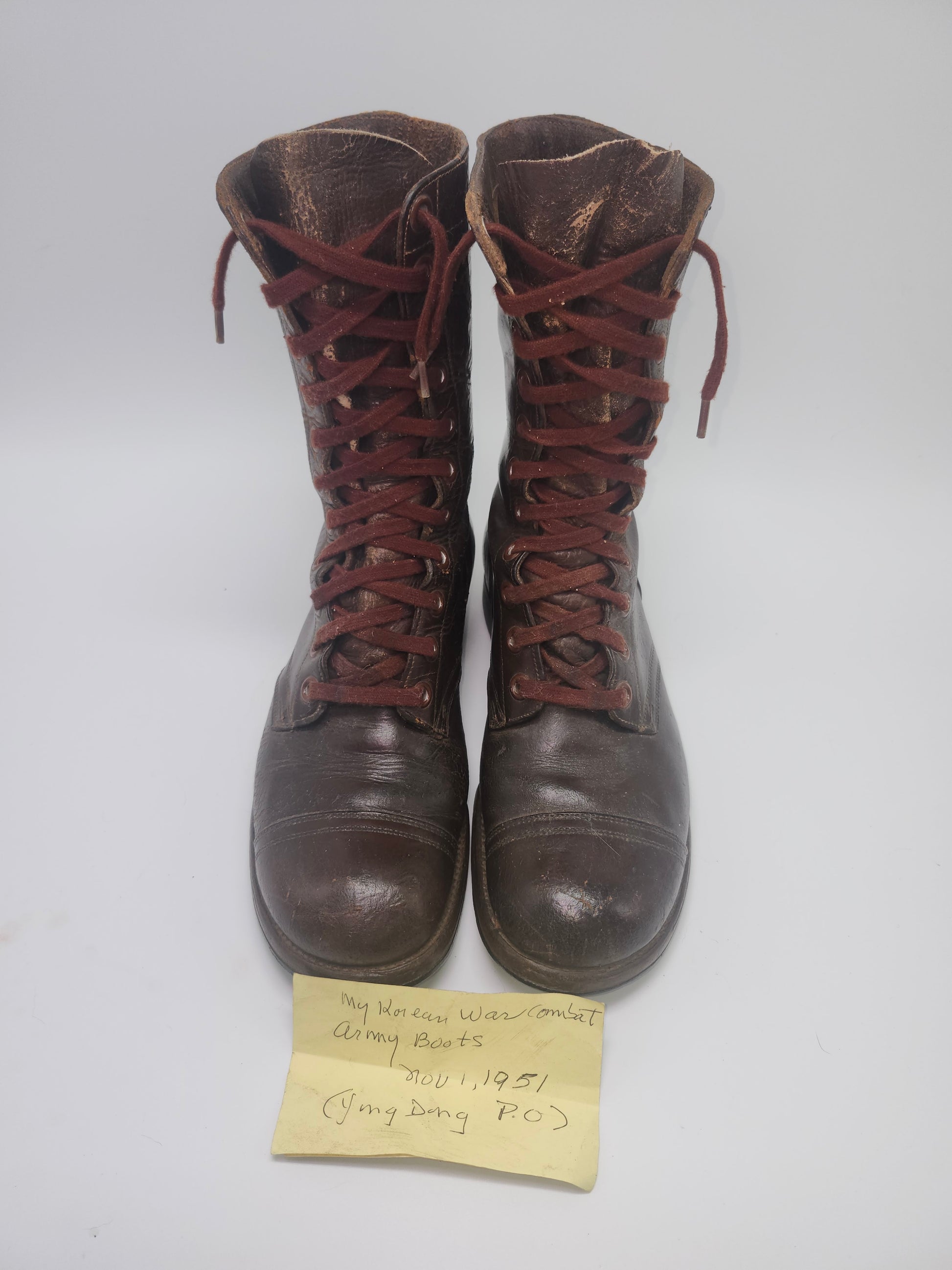 Pair of brown leather boots with red laces on a white background, accompanied by a yellow label reading, "my Korean war combat boots. Nov 1, 1951. Ying Dong, P.O."